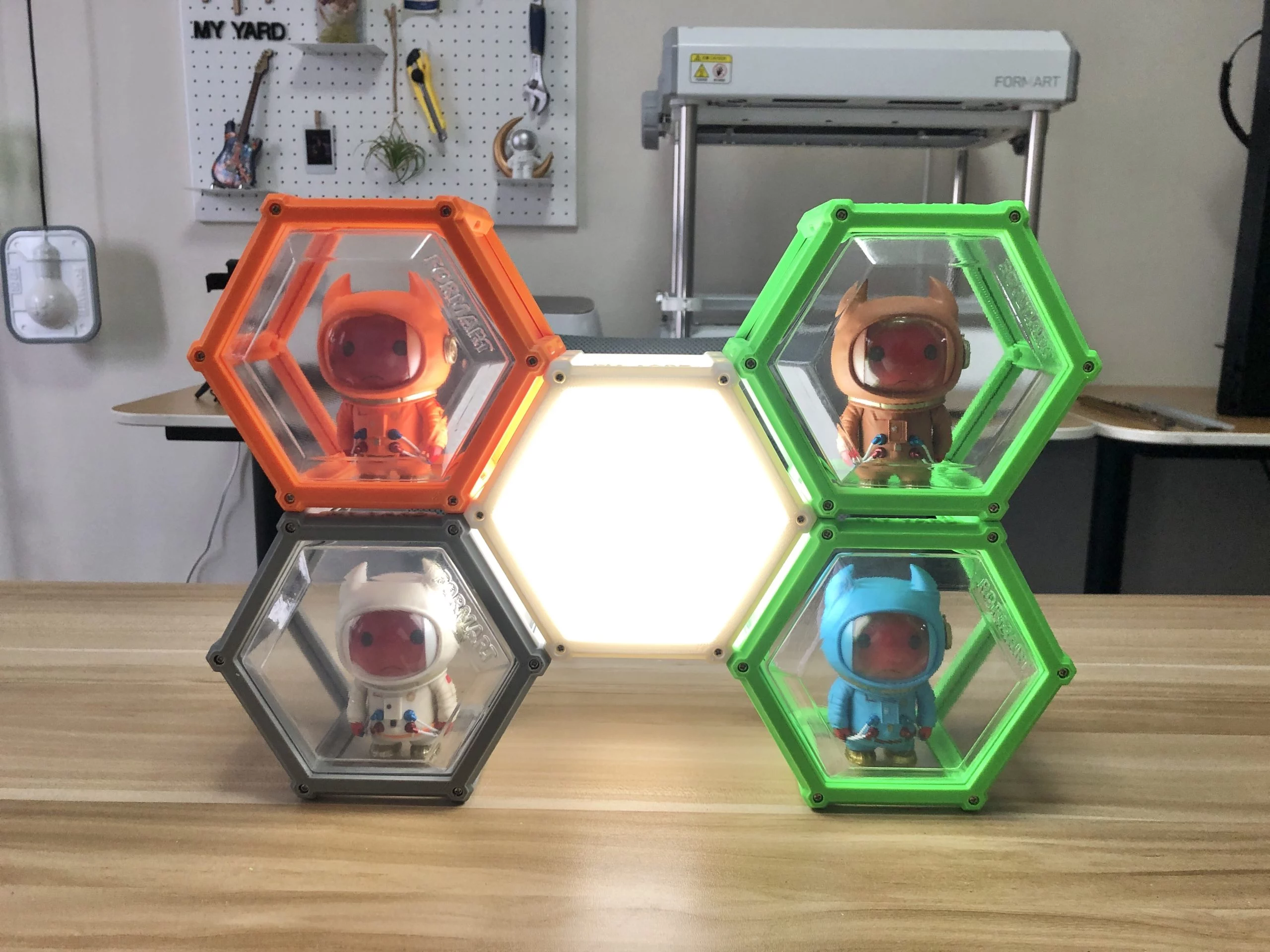 Transparent hexagonal display boxes with 3D-printed frames, stacked using magnetic design, containing small colorful figurines. The central hexagon frame features a white LED light.