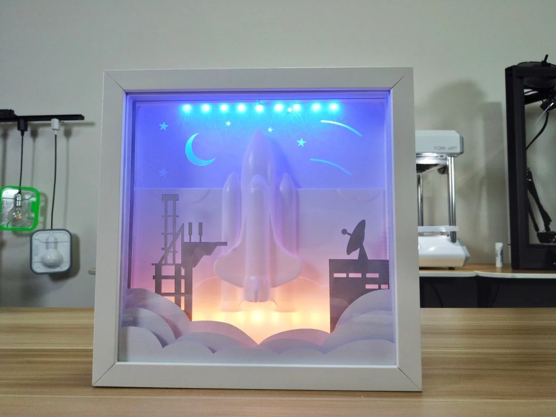A white frame contains a white space shuttle model in a launch position with flames, set against a blue starry background and accompanied by a gray launch pad. The back panel is illuminated with blue and red LED lights.