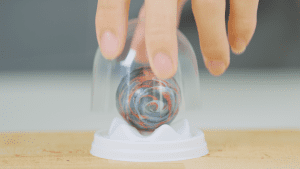 A hand gently placing a transparent cover over a finished chocolate ball, with a white display stand beneath