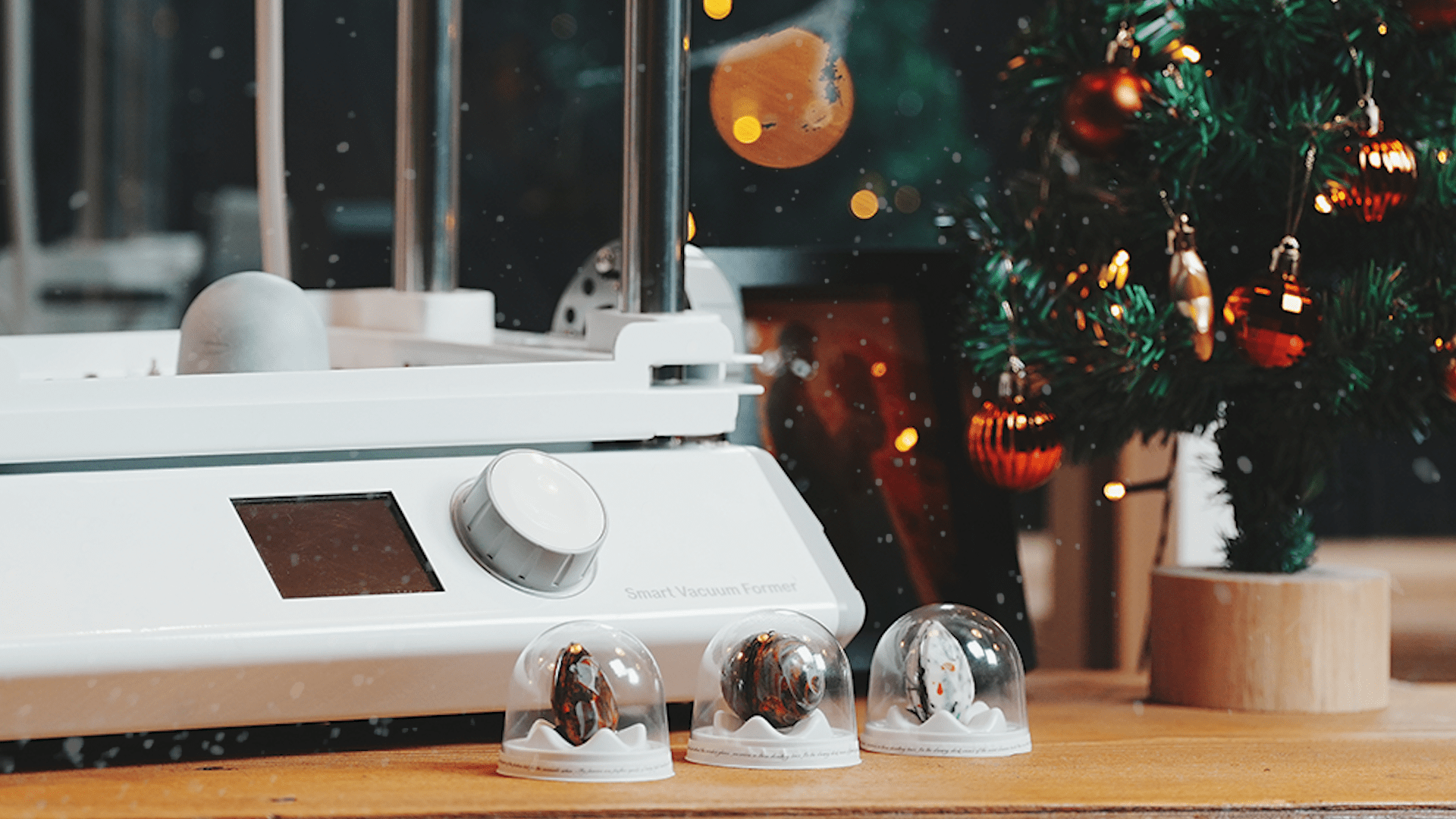 FORMART Smart Vacuum Former next to three finished chocolate molds placed on a white display stand, with a beautifully decorated Christmas tree in the background and snowflakes falling