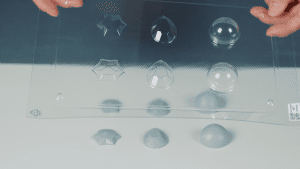 A hand holding a vacuum forming mold, showcasing finished chocolate molds with different shaped chocolate pieces at the bottom