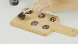 A hand using a stick to fill chocolate into a molded chocolate shell, with different shaped chocolate pieces placed on a wooden cutting board
