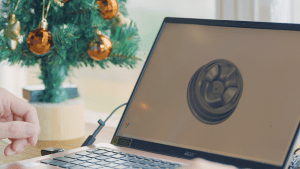 A hand operating a laptop, with the screen displaying a 3D mold design, and a Christmas tree with decorations in the background