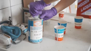 A worker wearing purple gloves is opening a paint can, preparing to apply paint. Nearby, there are paper cups and protective gear, with various paints and materials on the table.
