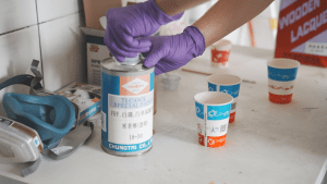 A worker wearing purple gloves is opening a paint can, preparing to apply paint. Nearby, there are paper cups and protective gear, with various paints and materials on the table.