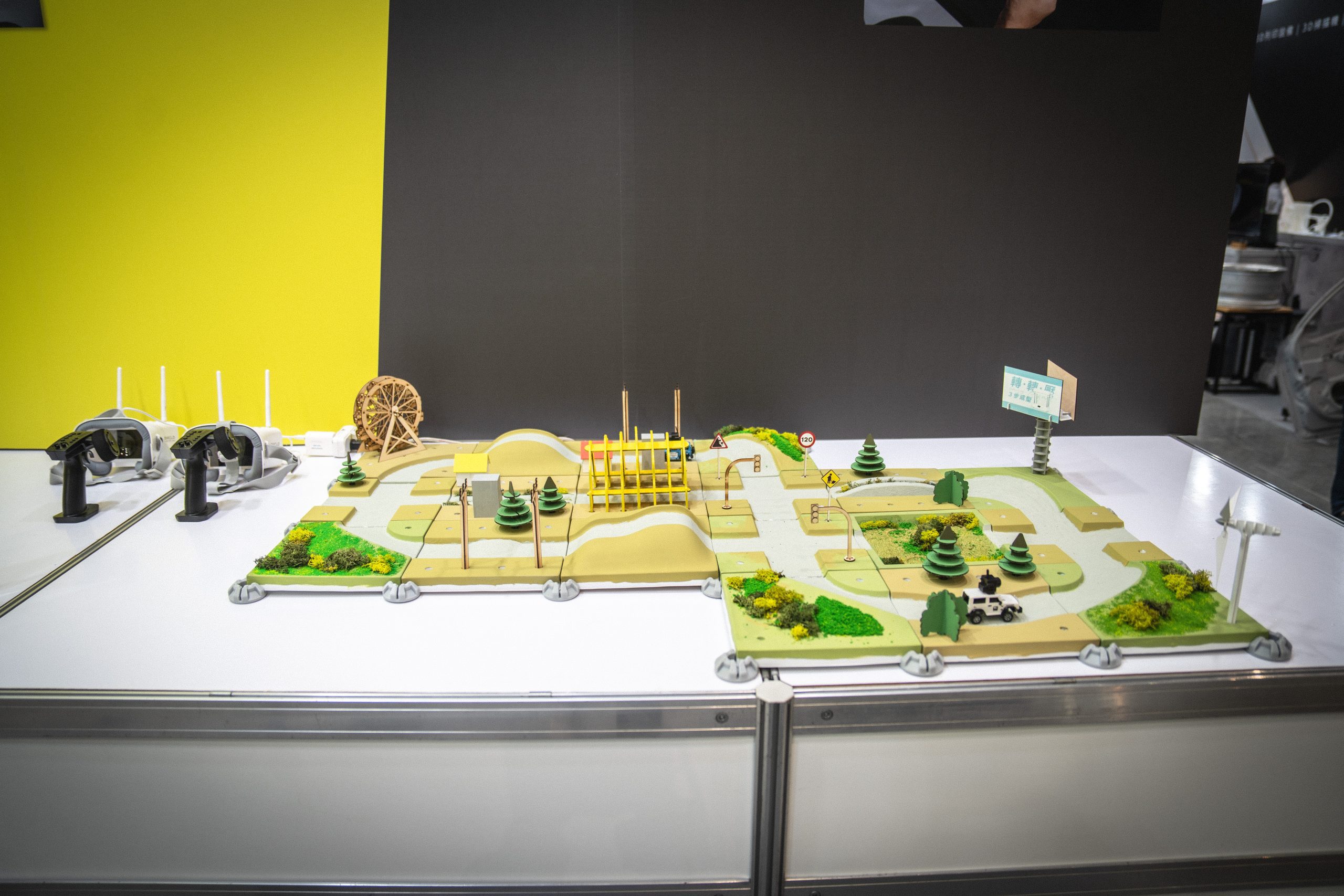 A display of an off-road track model surrounded by traffic signs, trees, buildings, and wind turbines. On the left, two controllers are shown, indicating the scene is being operated