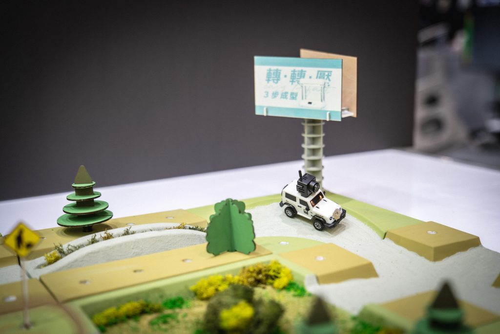 A small off-road car is driving on a simulated off-road track, with trees, traffic signs, and a large billboard in the background.