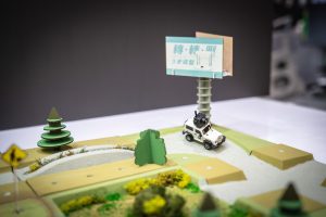 小型のオフロードカーがシミュレートされたオフロードトラックを走行しており、背景には木々、交通標識、大きな看板があります。