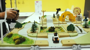 An operator is controlling a small RC car, with a background featuring a model off-road track made up of simulated roads, traffic signs, trees, and a tunnel.