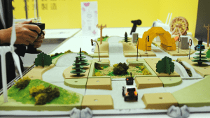 An operator is controlling a small RC car, with a background featuring a model off-road track made up of simulated roads, traffic signs, trees, and a tunnel.