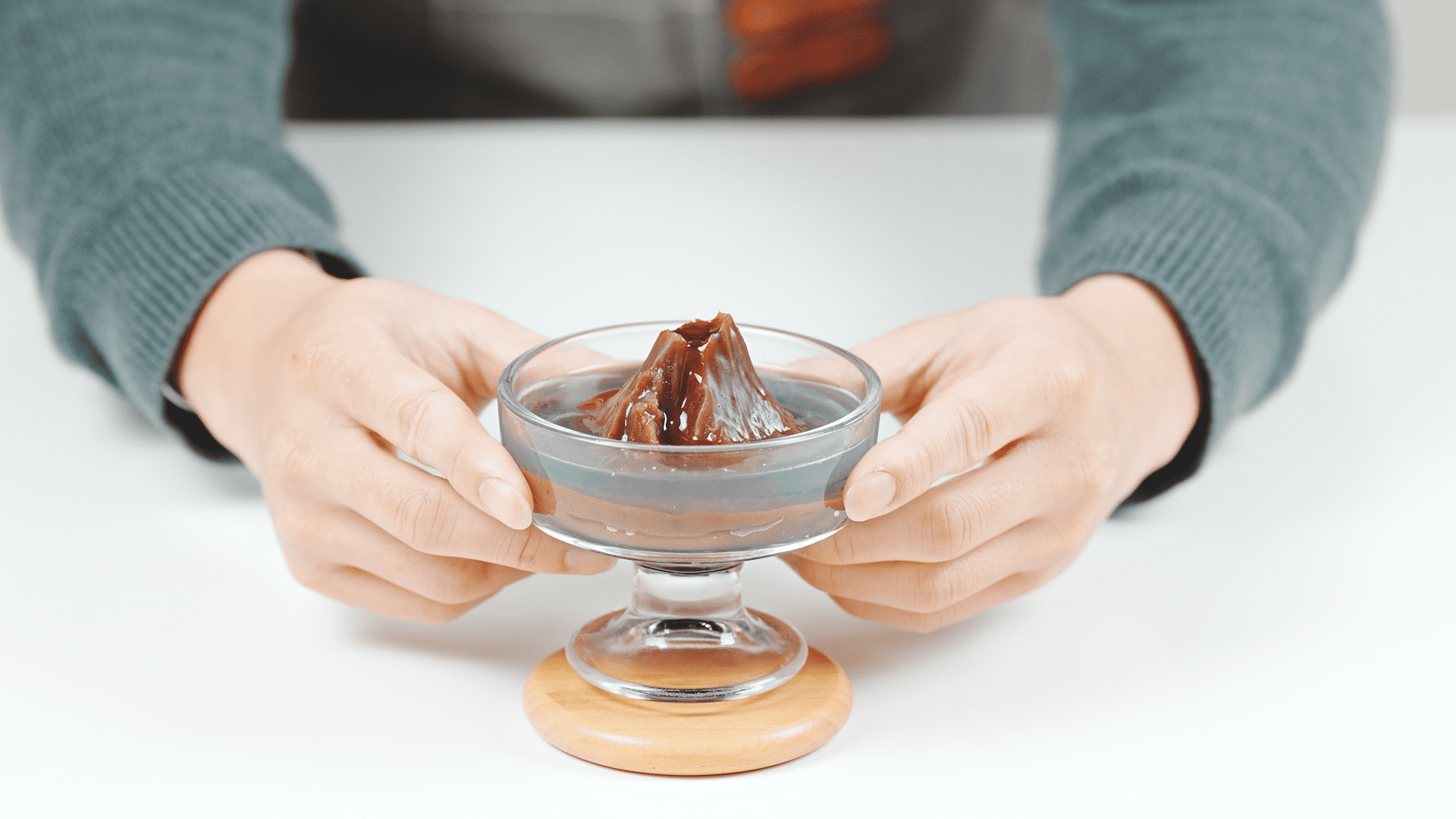 Hands holding a glass bowl with 'Snake Dance Red Rock Yokan' dessert, featuring a volcano-shaped yokan with layers of dark and light brown jelly.